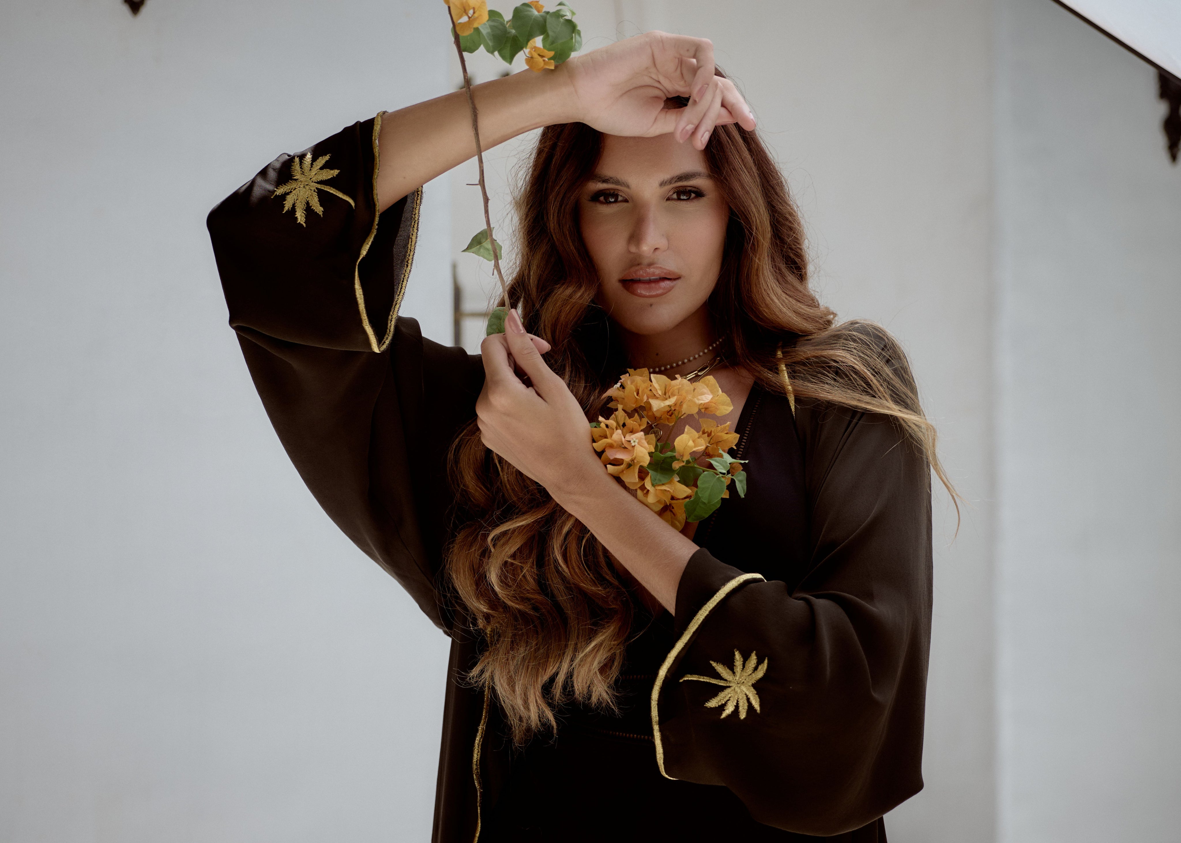 Woman wearing a black outfit with gold embroidery, holding a plant against a white background