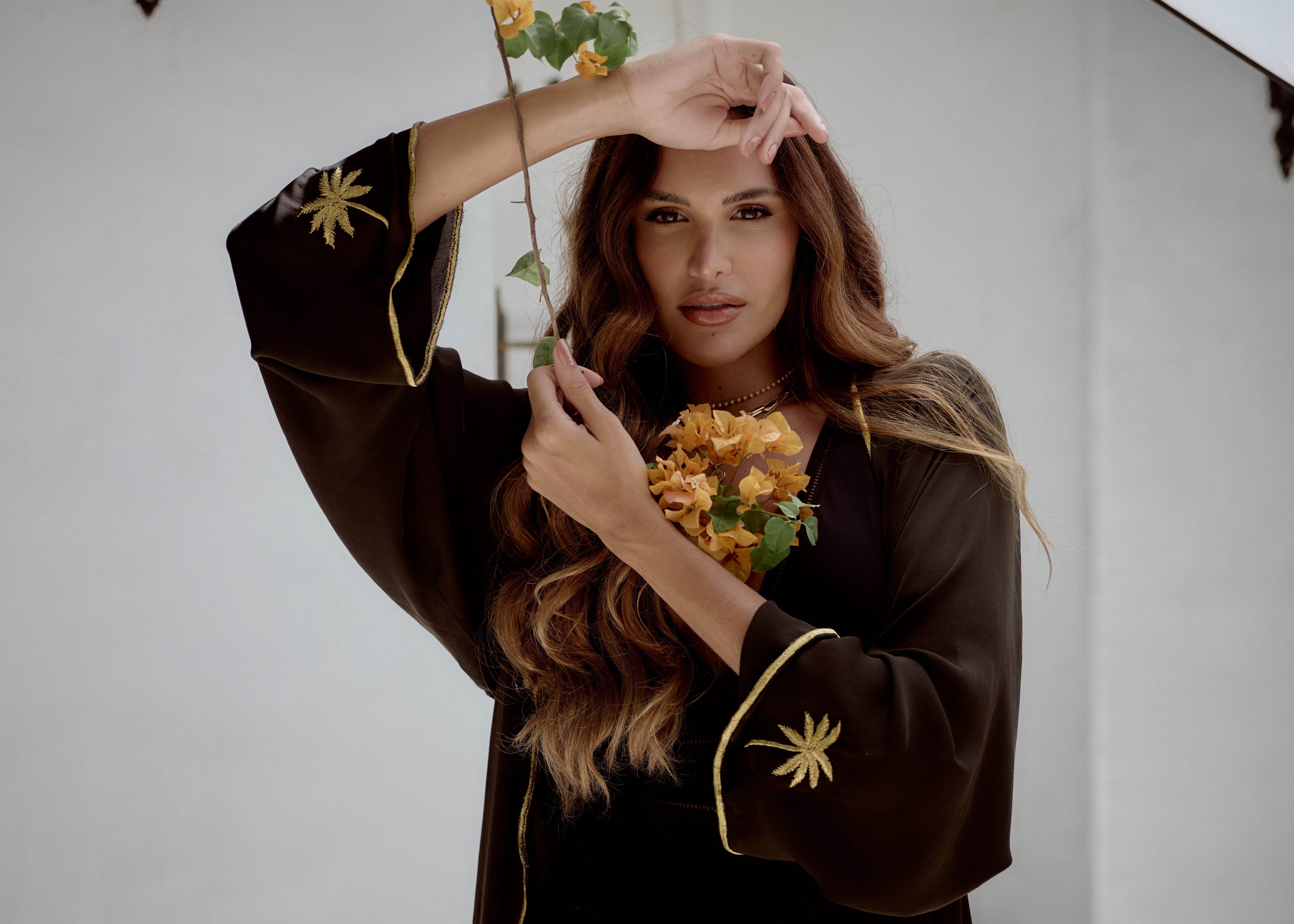 Woman wearing a black outfit with gold embroidery, holding a plant against a white background