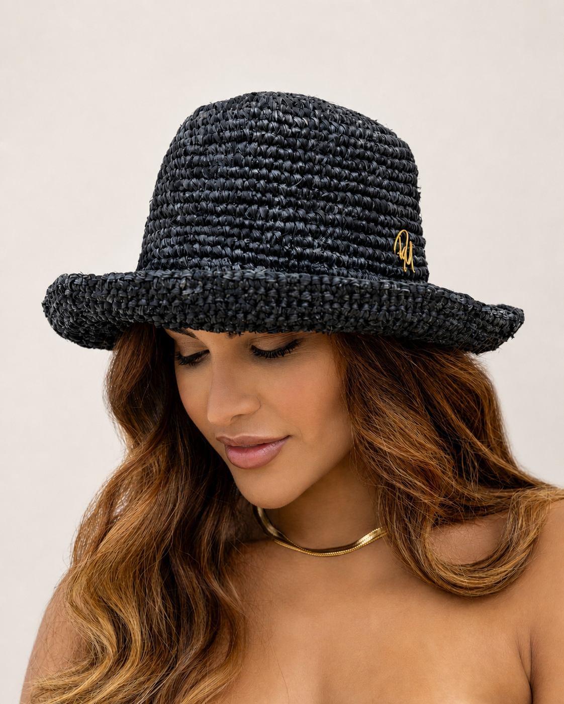 Woman wearing a black woven hat with a gold emblem on a beige background