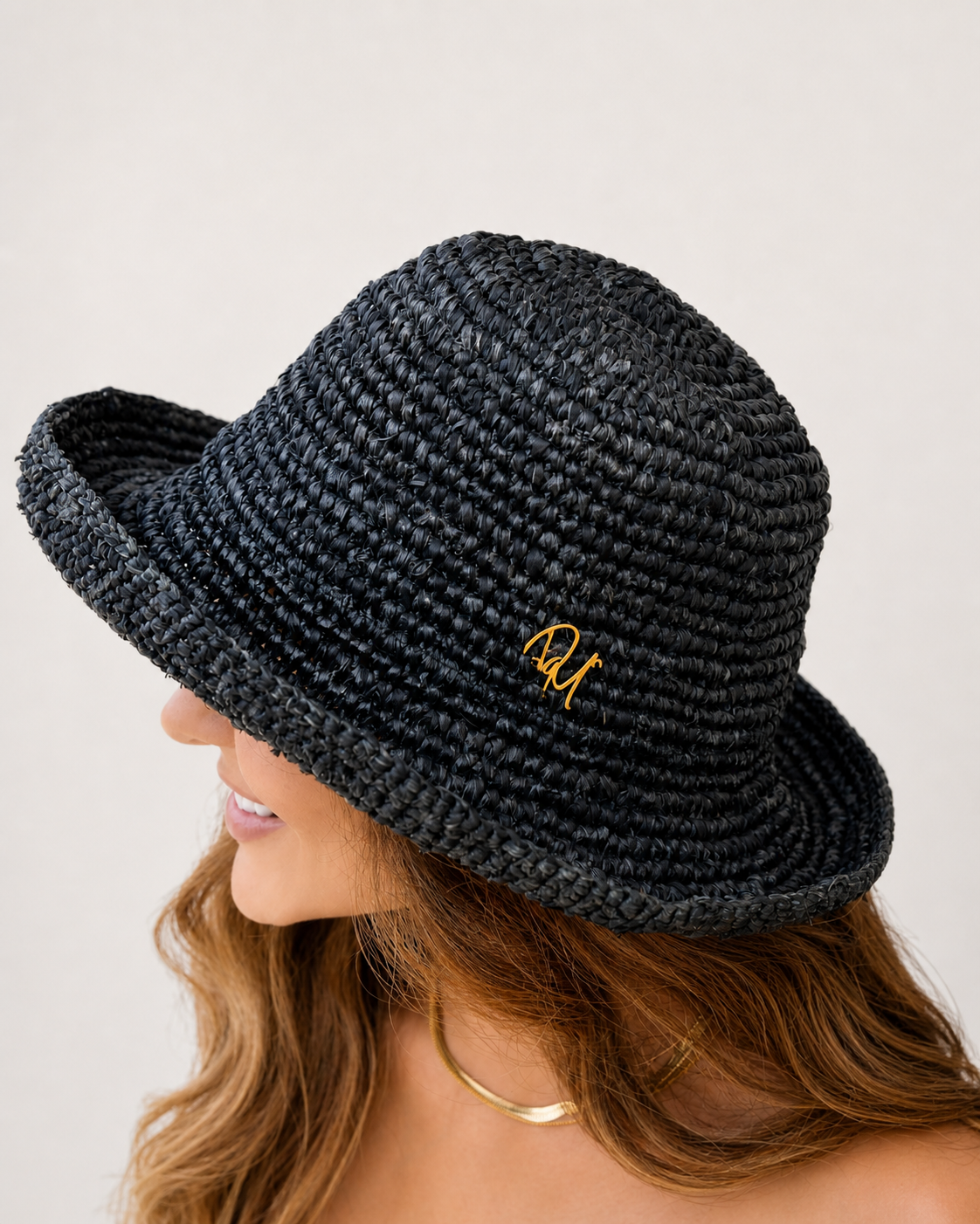 Black woven hat with a small gold logo worn by a person on a light gray background